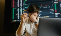 A stressed man looks at stock market data on his computer screen in an office setting.